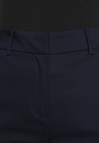 Marina byxor med en slät tygkvalitet. Har en platt front, sidofickor och subtila sömnadsdetaljer. En del av midjebandet är synligt ovanför.