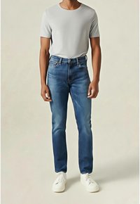 Homme portant un t-shirt à manches courtes gris clair, un jean slim bleu et des baskets blanches, debout devant un fond beige uni.