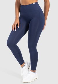 Marineblauwe high-waisted leggings van rekbare stof, met een gladde textuur en naaddetails, gecombineerd met witte sport sneakers.