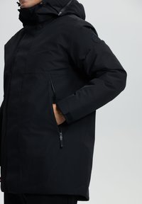 Schwarze wasserfeste Jacke mit Kapuze, ausgestattet mit Reißverschlusstaschen, verstellbaren Manschetten und einer glatten Textur. Einfaches Design ohne sichtbare Muster.