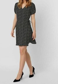 Schwarzes Kleid mit weißen Pünktchen, V-Ausschnitt, kurzen Puffärmeln, tailliertem Schnitt und ausgestelltem Rock, kombiniert mit schwarzen High Heels.