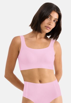 Femme aux cheveux bruns mi-longs portant un crop top rose clair sans coutures et une culotte taille haute, sur un fond uni.