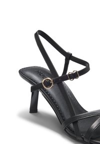 Μαύρο δερμάτινο πέδιλο με ψηλό τακούνι, λεπτά λουράκια που διασταυρώνονται, δύο αγκράφες σε χρυσό τόνο και κωνικό τακούνι.