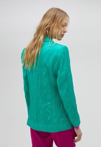 Blazer de textura turquesa con un acabado arrugado, que presenta un cierre de un botón y una silueta ajustada, combinado con pantalones de color rosa brillante.