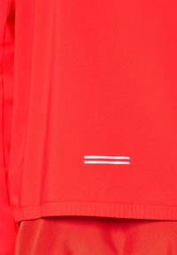 Chemise de sport rouge avec de petites rayures horizontales doubles blanches près de l'ourlet, présentée avec un pantalon rouge assorti.