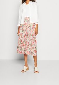 Camicia bianca a bottoni sopra una maglia bianca, abbinata a una gonna midi floreale nei toni di rosa, verde e beige. Indossa sandali aperti di colore avorio.