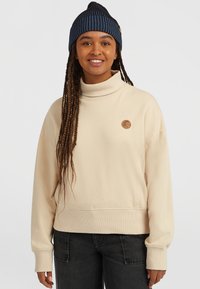 Beige sweatshirt med hög krage, ribbade manschetter och en texturerad logotypapplikation. Kombinerad med mörka jeans och en svart och blå randig mössa.