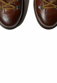 Botas de senderismo de cuero marrón con cordones amarillos, ojales de metal, puntera redondeada y suela texturizada, vistas desde arriba sobre un fondo blanco.