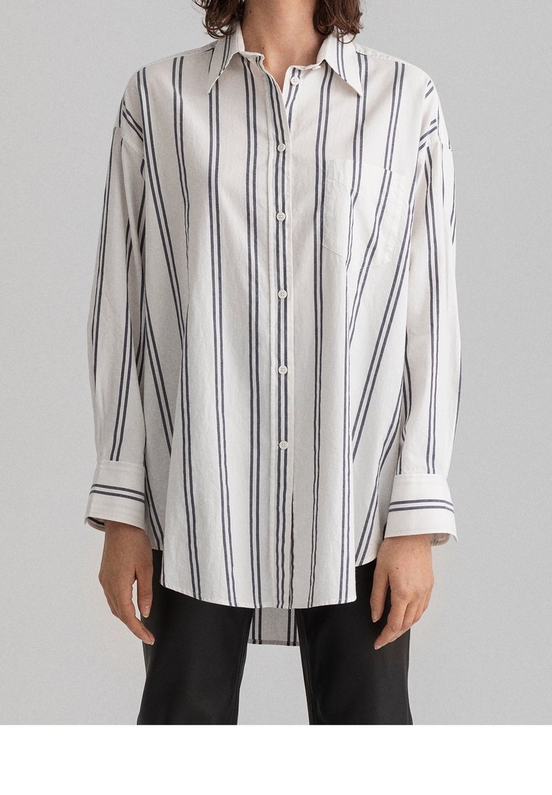 Personne portant une chemise blanche à rayures noires verticales avec une poche poitrine devant, associée à un pantalon noir.