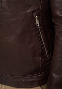 Donkerbruine leren jas met een gestructureerd oppervlak, voorzien van een zijritszak en een getailleerde pasvorm. Zichtbare stiksels langs de naden.