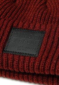 Burgundi kötött sapka bordázott textúrával, fekete bőr emblémával, amelyen a "MINOTI SURPLUS APPAREL GOODS EST. 1957." felirat található.