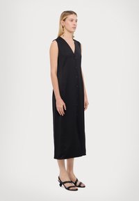 Rochie neagră fără mâneci, cu decolteu în V și butoane pe față. Material neted, cu o siluetă fluidă, care ajunge până la jumătatea gambei, asortată cu sandale negre.