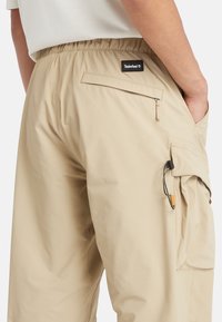 Beige cargobyxor med elastisk midja, sidoficka för cargo och bakficka med dragkedja. Strukturerad tyg med lätt design.