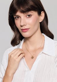 Kobieta w białej bluzce trzyma złoty naszyjnik z zawieszką w kształcie drzewa, prezentując delikatny, naturalny makijaż na neutralnym tle.