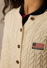 Kremasti kardigan s pletenicom i okruglim ovratnikom, s smešnim dugmadi i zakrpom s američkom zastavom i logom "SUPERDRY".
