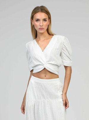 Femme aux cheveux blonds raides portant un chemisier court blanc à manches bouffantes et une jupe taille haute assortie sur fond gris.