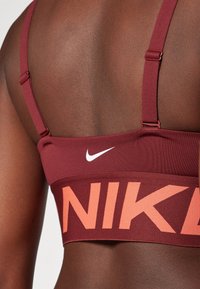 Bordo športni modrček z nastavljivimi naramnicami, ki vsebuje velik oranžen logotip "NIKE" in majhen bel tek na hrbtni strani. Gladka tekstura.