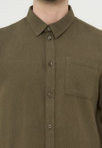 Κοντινό πλάνο ενός ατόμου που φοράει πουκάμισο σε ελιά πράσινο με κουμπιά, μυτερό γιακά και τσέπη στο αριστερό μέρος του στήθους.