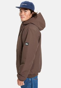 Ung man iklädd en brun huva-jacka, blå Quiksilver-keps och blå jeans står mot en helt vit bakgrund.