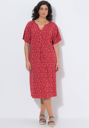 Donna in piedi che indossa un vestito rosso al ginocchio con stampa astratta blu, davanti con bottoni, maniche corte e sandali con plateau blu.