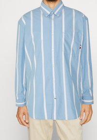 Camisa de manga larga azul con botones y rayas verticales blancas, bolsillo en el pecho izquierdo y pequeño detalle de logo en la manga. Tejido de algodón.