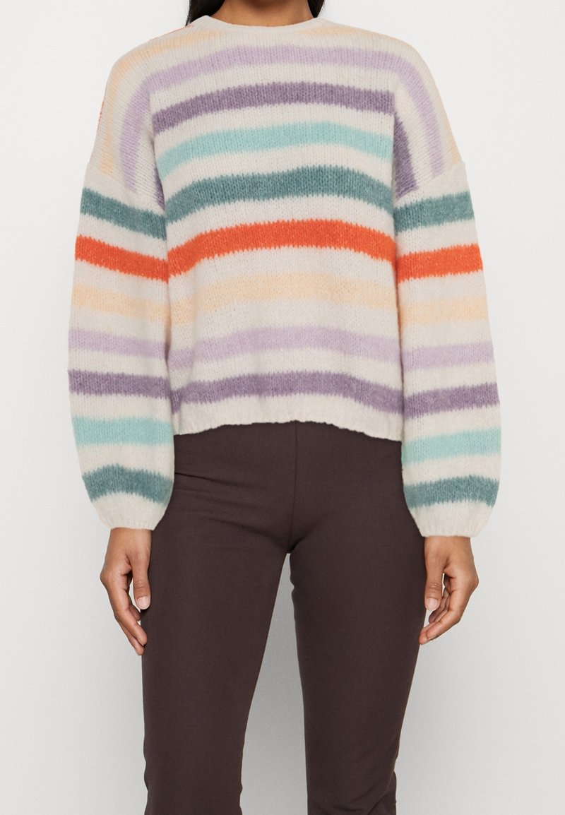 Pull duveteux à rayures horizontales violet, sarcelle, orange et beige, porté avec un pantalon marron foncé, montrant le haut du torse et les bras.