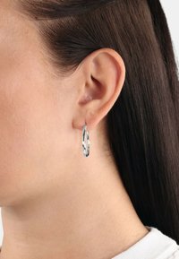 Pendiente de aro de plata con un diseño retorcido, que presenta una superficie brillante y reflectante. Adecuado para orejas perforadas, colocado en el lóbulo de la modelo.