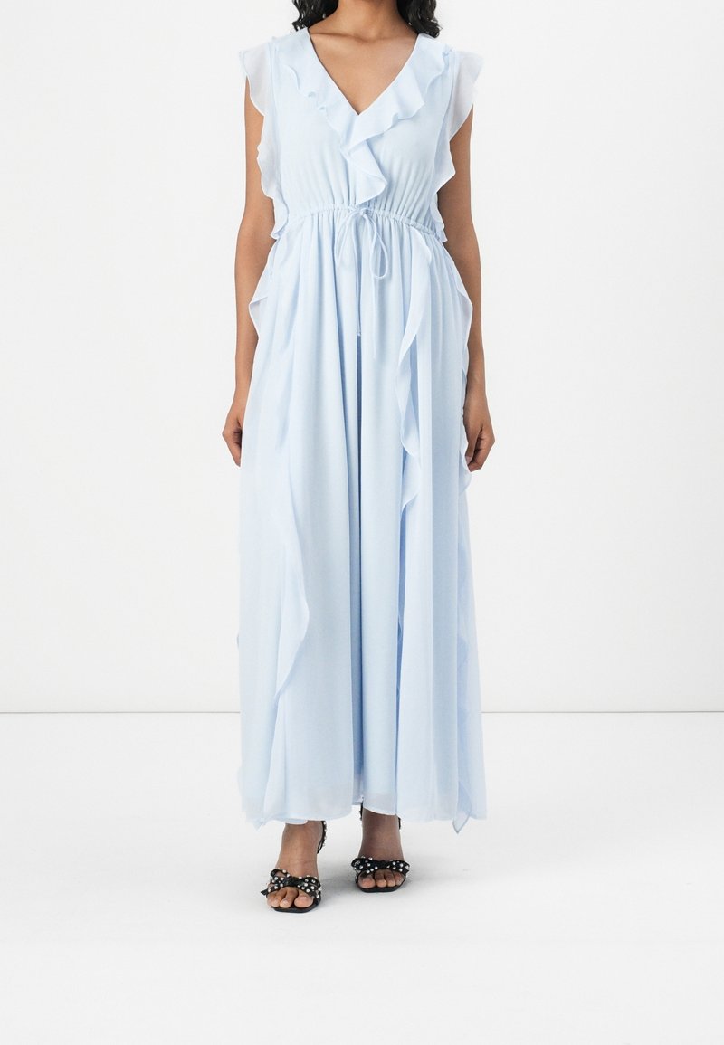 Femme portant une robe longue bleu clair sans manches avec des détails à volants et une taille cintrée, associée à des sandales noires à pois.