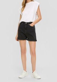 Witte t-shirt met korte mouwen, hoge zwarte denim shorts met opgerolde manchetten, en witte sneakers, staande op een effen achtergrond.