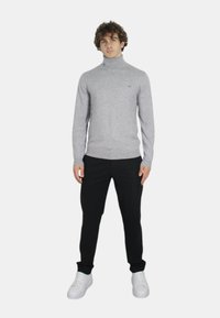 Maglione a collo alto grigio chiaro realizzato in tessuto lavorato a maglia, design aderente, abbinato a pantaloni neri e sneakers bianche, in piedi su uno sfondo neutro.
