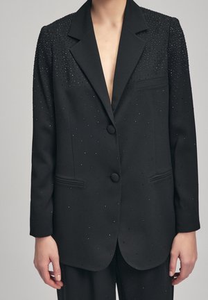 Zwarte op maat gemaakte blazer met kleine fonkelende versieringen, twee knopen en klepzakken, gedragen over bijpassende broek door een persoon.