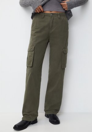 Persoon draagt olijfgroene cargobroek, zwarte laarzen en een grijze trui, handen passen de zoom van de trui boven de taille aan.