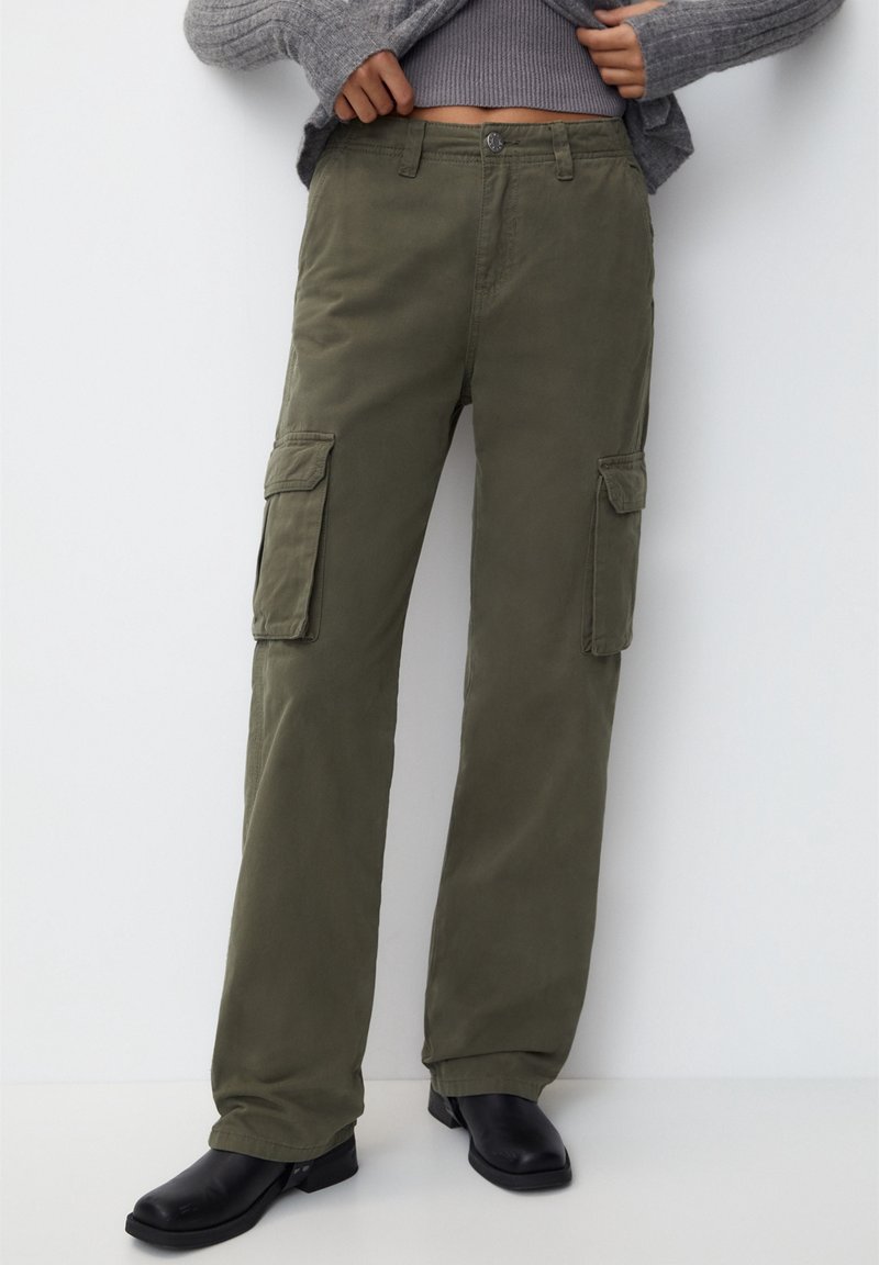 Personne portant un pantalon cargo vert olive, des bottes noires et un pull gris, mains ajustant l'ourlet du pull au-dessus de la taille.