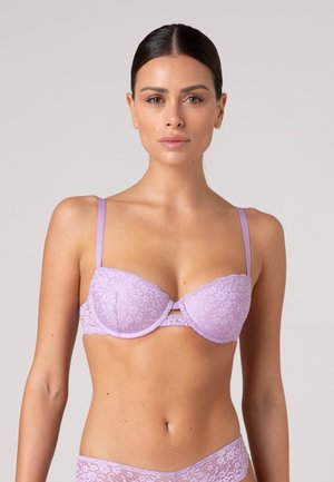 Donna con capelli scuri raccolti, indossa un reggiseno in pizzo lavanda e mutandine in pizzo abbinate, in piedi davanti a uno sfondo chiaro e semplice.