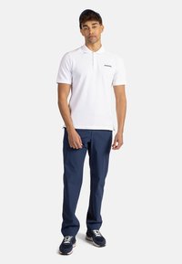 Valkoinen polo-paita lyhyillä hihoilla ja kauluksella, yhdistettynä tummansinisiin housuihin ja sinisiin urheilukengiin. Yksinkertainen muotoilu, ei kuvioita tai koristeita.