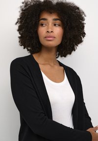 Crna kardigan od mekog materijala, otvorenog prednjeg dijela, dugih rukava, u kombinaciji s jednostavnim bijelim T-shirtom, na neutralnoj pozadini.