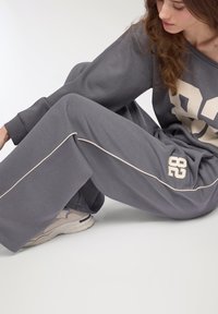 Kobieta siedząca na podłodze, ubrana w szare spodnie dresowe z białym paskiem, pasującą bluzę z nadrukiem "82" oraz off-white sneakersy.