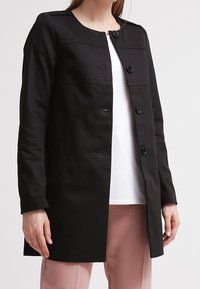 Nainen, jolla on päällään musta napillinen takki valkoisen paidan ja vaaleanpunaisia housujen päällä, seisoo vaaleaa taustaa vasten.