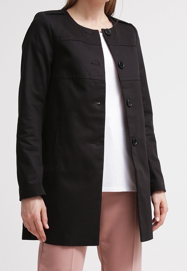 Nainen, jolla on päällään musta napillinen takki valkoisen paidan ja vaaleanpunaisia housujen päällä, seisoo vaaleaa taustaa vasten.