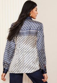 Seidene Bluse mit einem navyblauen und weißen Muster aus verlängerten Ovale; lange Ärmel, knöpfbare Manschetten und eine glatte Textur.