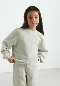 Felpa grigia heather con maniche lunghe e scollo rotondo, abbinata a pantaloni della tuta coordinati. Piccolo dettaglio del logo giallo sul petto.