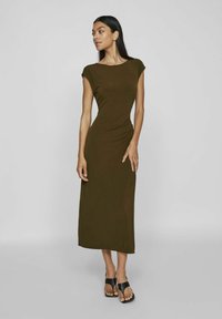 Olivovo zelená, priliehavá maxi šaty s kapucňovými rukávmi a zhromaždeným detailom na boku. Nosené s čiernymi sandálmi na viazanie na neutrálnom pozadí.