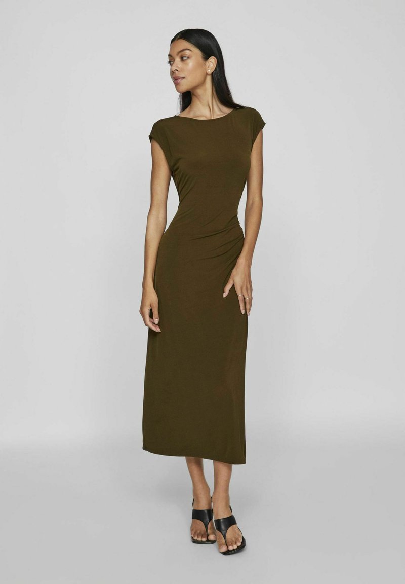 Olivovo zelená, priliehavá maxi šaty s kapucňovými rukávmi a zhromaždeným detailom na boku. Nosené s čiernymi sandálmi na viazanie na neutrálnom pozadí.