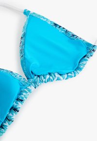 Top de bikini turquesa con una tela suave, copas triangulares y un borde texturizado azul y blanco. Incluye tirantes ajustables.