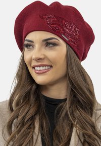 Béret tricoté rouge en tissu texturé avec un motif de strass scintillants. Coupe douce et loose avec une forme arrondie.