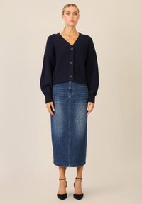 Marineblau gerippter Cardigan mit Knöpfen, kombiniert mit einem knielangen Jeansrock mit gepunktetem Muster und taillierter Silhouette. Schwarze High Heels.