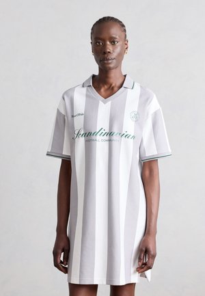 Mustanahaline naine kannab lahtist halli ja valget vertikaaltriibulist jalgpallistiilis kleiti, millel on ees tekst „Scandinavian Football Community“.