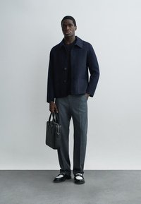 Homme portant une veste bleu marine foncé, un pantalon gris, des mocassins noir et blanc, tenant une mallette en cuir noire sur un fond uni.