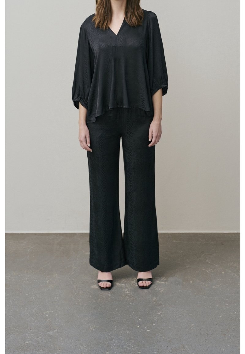 Femme portant une blouse noire ample à manches trois-quarts, un pantalon large assorti et des sandales noires à bouts ouverts, debout sur un sol gris.