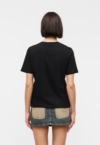 Personne avec des cheveux courts et foncés portant un t-shirt noir uni à manches courtes et une mini-jupe en jean avec des poches arrière beiges, de dos.